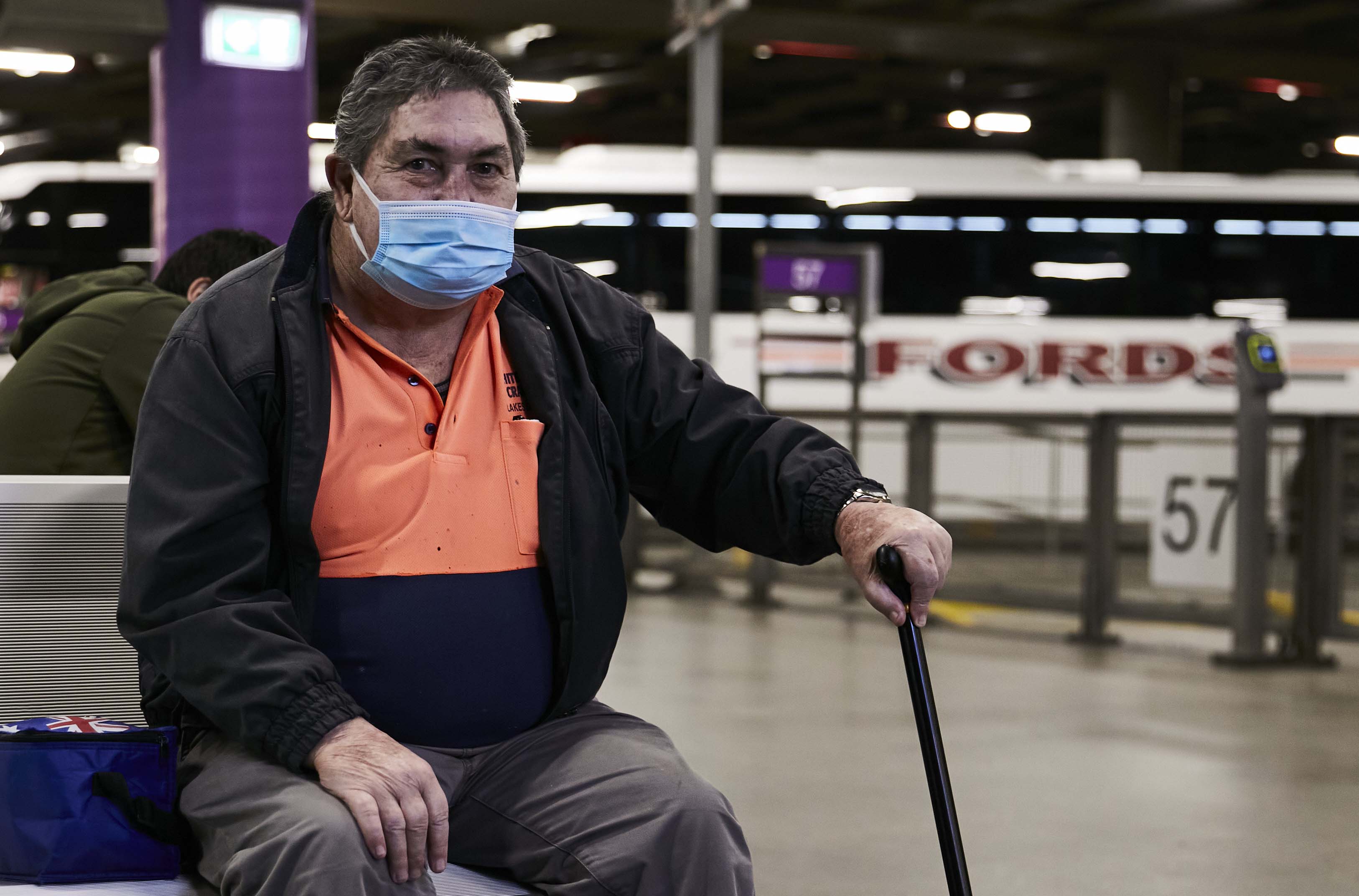 Image shows a middle-aged man sitting in a bus depot. He is wearing a face mask and a high-vis shirt. He has a walking stick in his left hand.