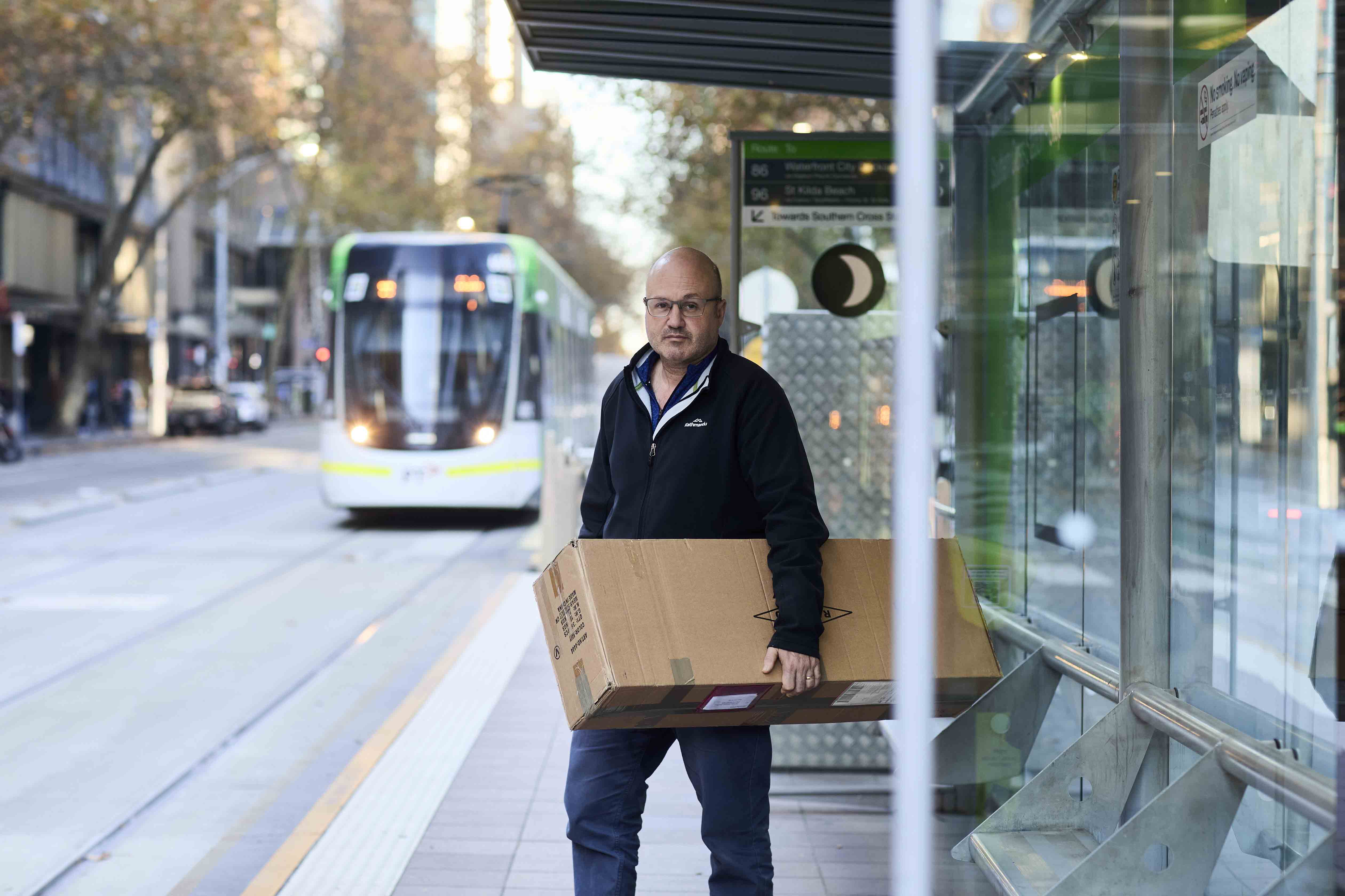 Image shows a middle-aged man waiting at a tram stop. He is carrying a large cardboard box. There is a tram approaching behind him.