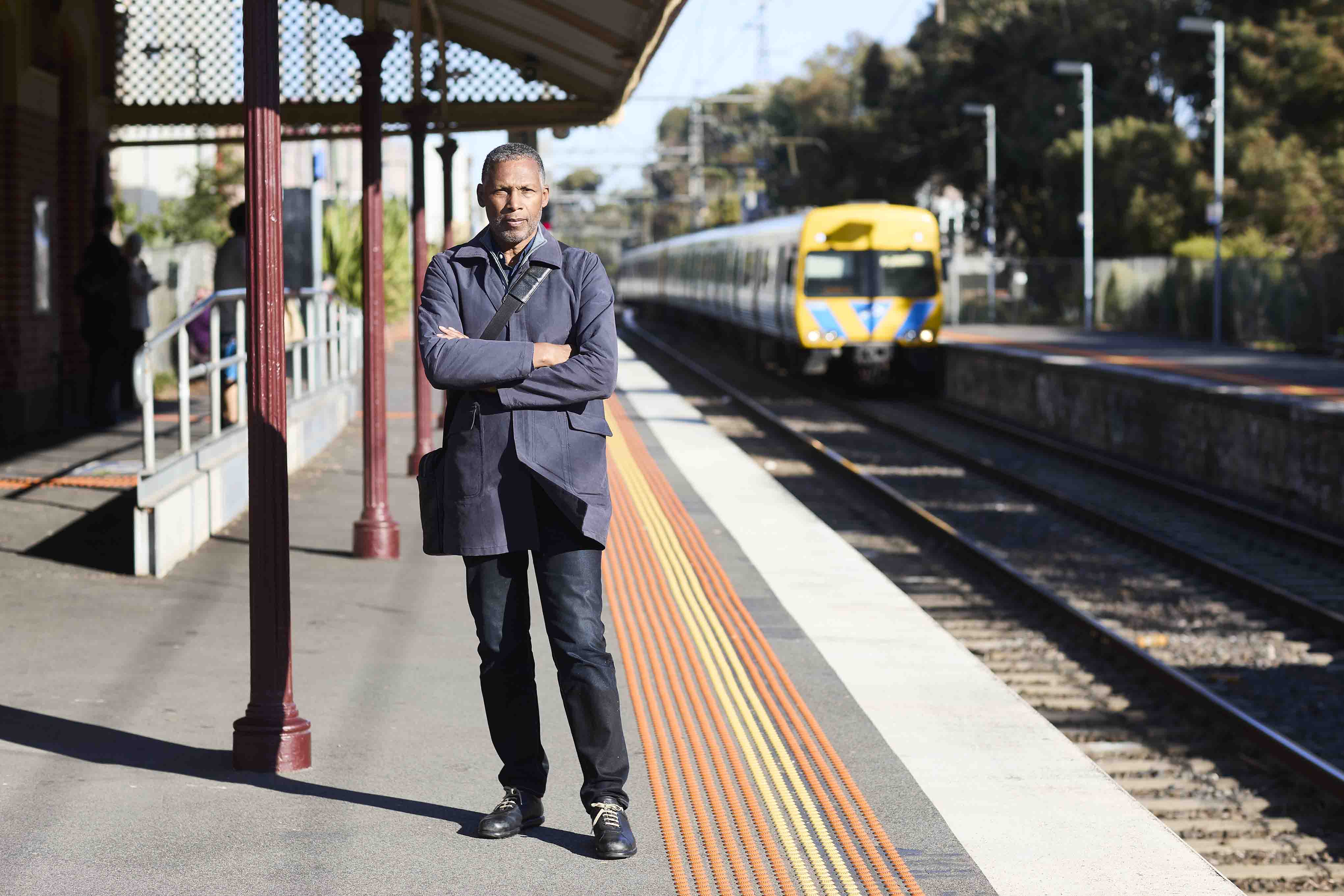 Image shows a man standing on a train platform with his arms crossed. There is a train approaching in the background.