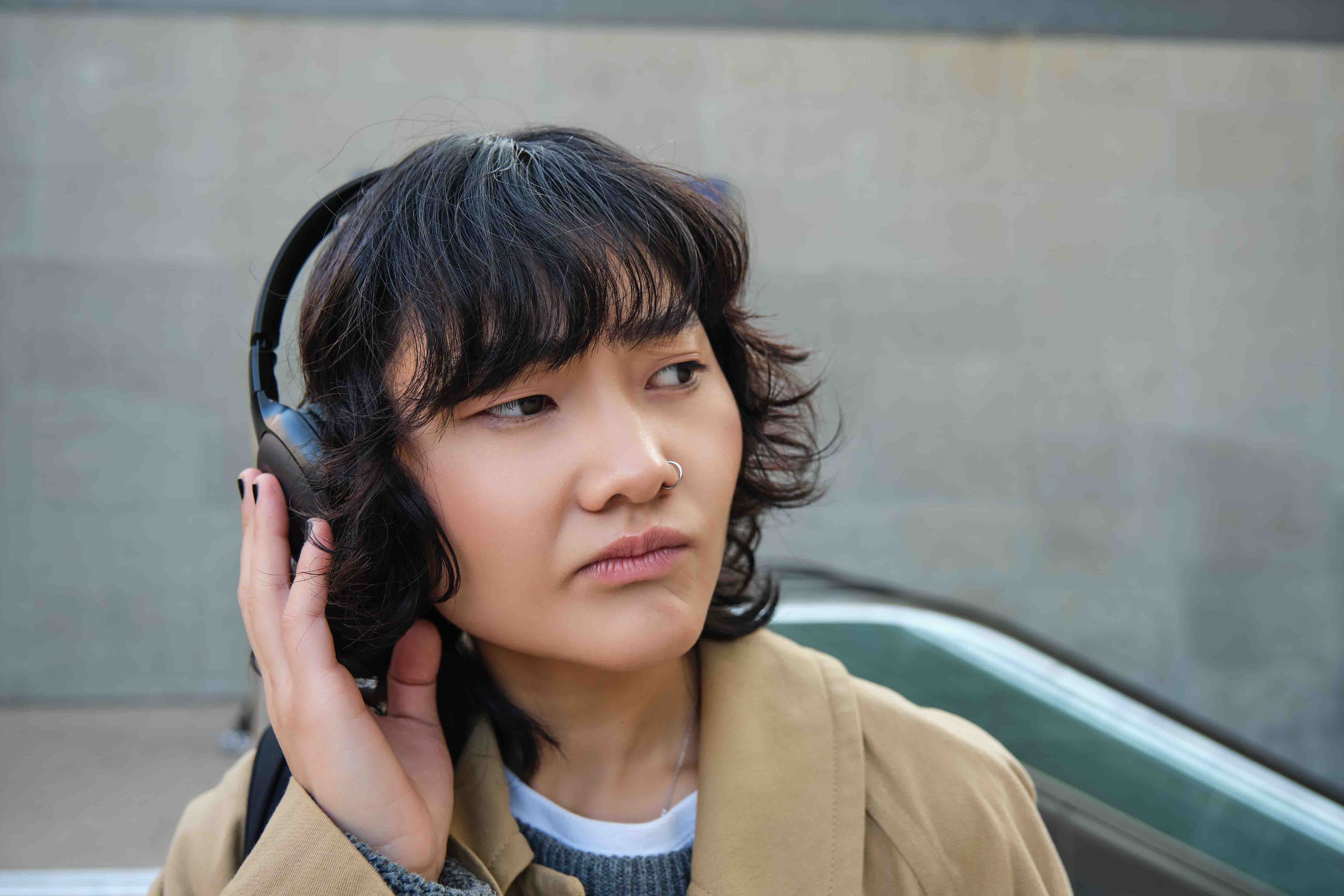 Image shows a young woman waiting for public transport.She is wearing headphones and looking to her left.