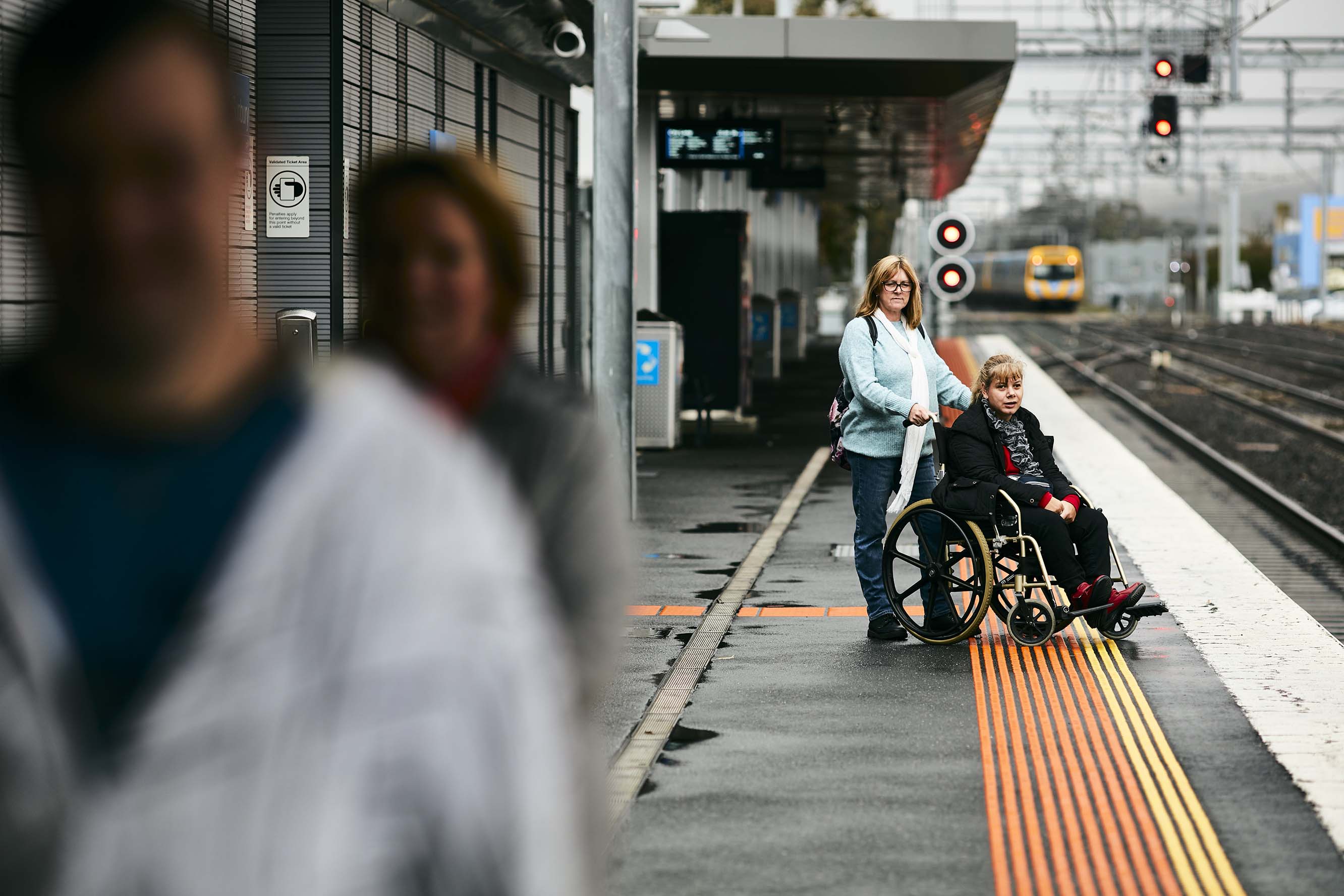 Image shows a person in a wheelchair waiting on a station platform with her carer as a train approaches in the background.