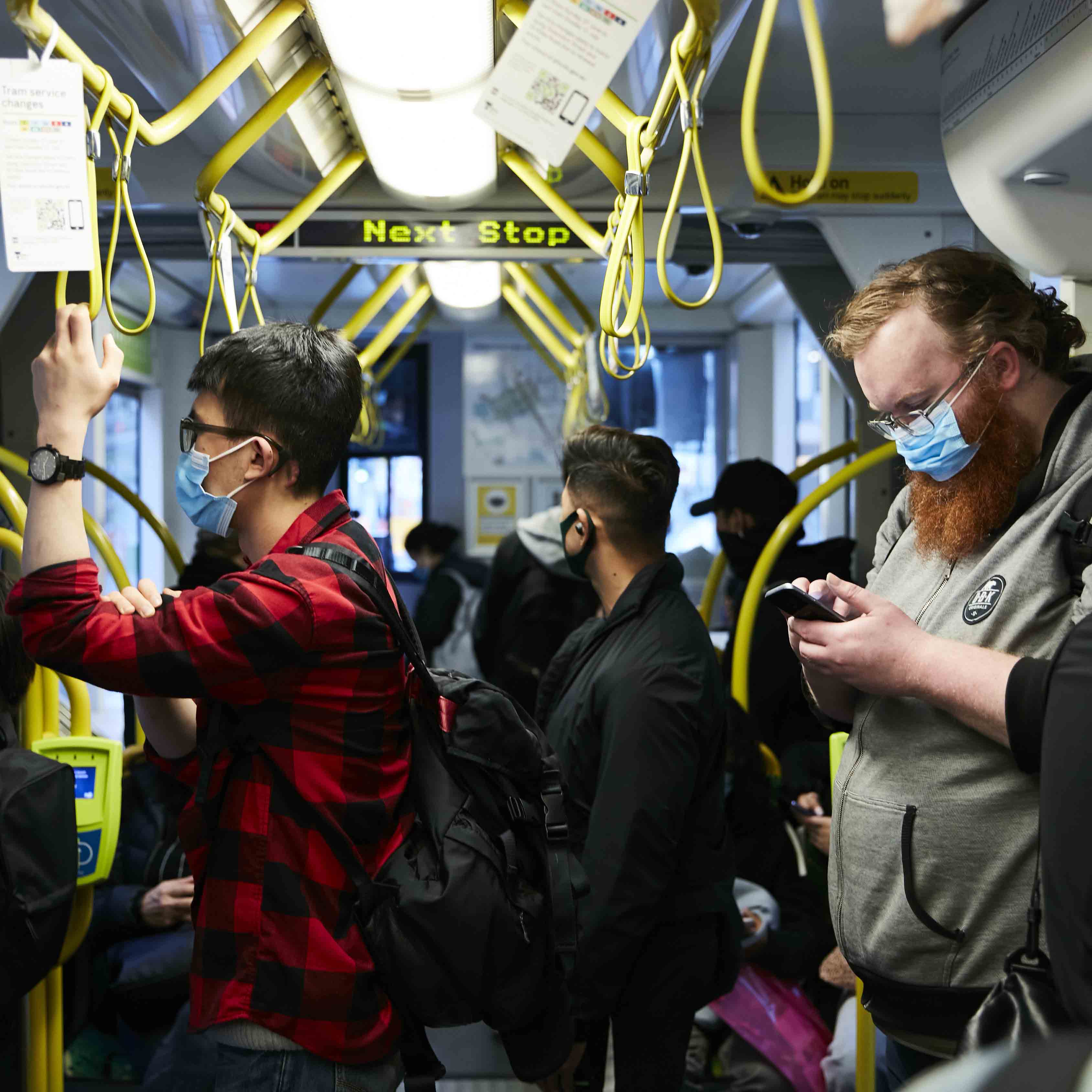 Image is a colour photograph showing passengers travelling on a tram. A person in a red and black checkered shirt stands to the left of the image holding onto a yellow hanging strap. They have short black hair and are wearing dark-framed glasses. A person standing to the right of the image has a red beard and medium-length red hair. They are wearing glasses and a grey hoodie and are looking down at the mobile phone in their hands. Several other people can be seen in the image – their backs to the camera. All of the passengers are wearing facemasks as they travel during the COVID-19 pandemic.