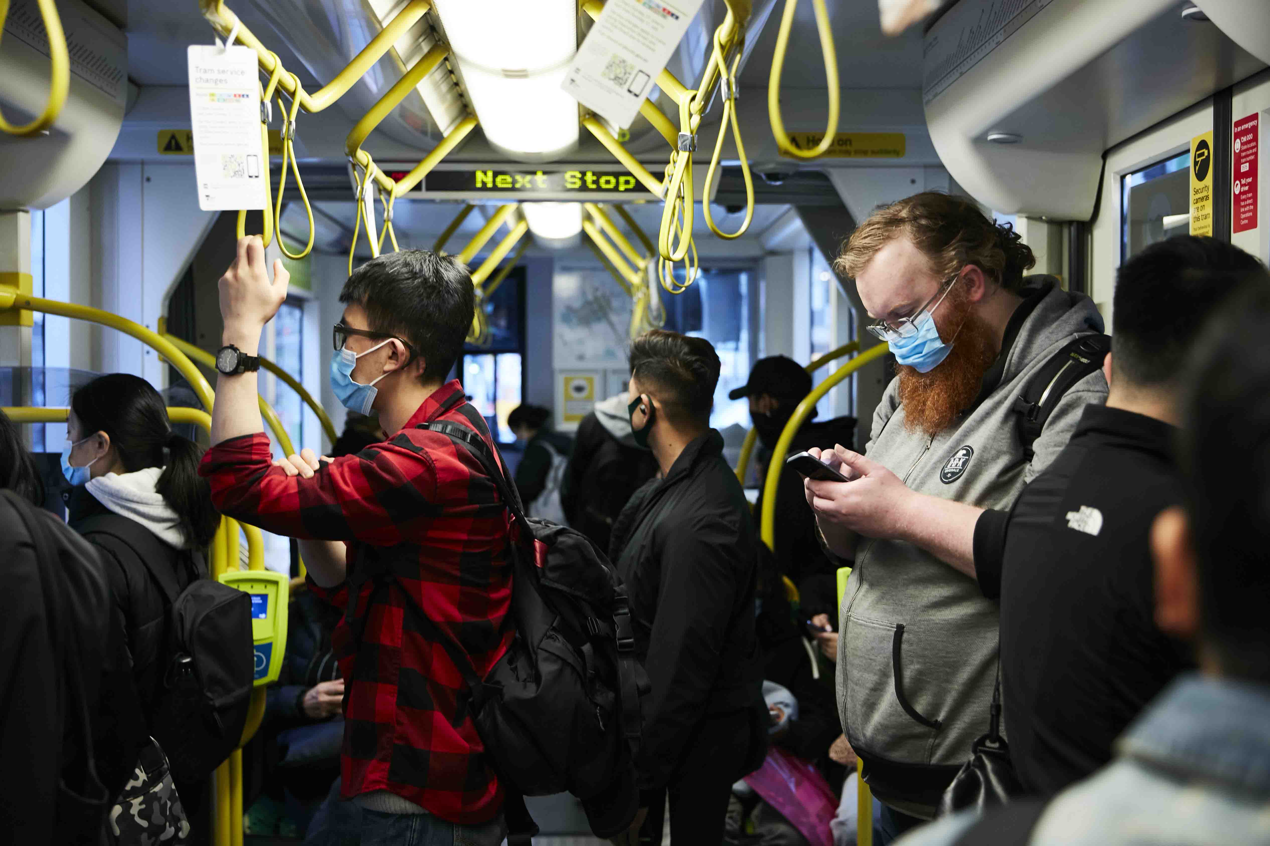 Image is a landscape orientation colour photograph showing passengers travelling on a tram. A person in a red and black checkered shirt stands to the left of the image holding onto a yellow hanging strap. They have short black hair and are wearing dark-framed glasses. A person standing to the right of the image has a red beard and medium-length red hair. They are wearing glasses and a grey hoodie and are looking down at the mobile phone in their hands. Several other people can be seen in the image – their backs to the camera. All of the passengers are wearing facemasks as they travel during the COVID-19 pandemic.